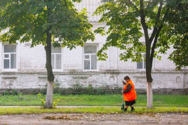 Beyaz tuğla bina ve ağaçların yakınındaki yaprakları süpüren turuncu yelekli belediye çalışanı. Sonbahar kentsel bakım sahnesi. Yayın ve toplum farkındalığı kampanyaları için tasarım.