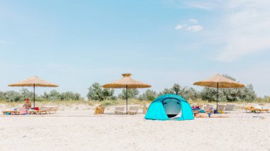 Kinburnska Kosa, Ukraine, August 16, 2017: A blue beach tent set up near straw umbrellas and sunbeds on a sandy shore under a clear blue sky. clipart