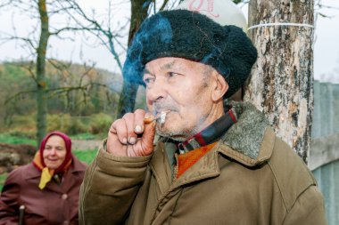 Geleneksel şapkalı yaşlı adam dışarıda sigara içiyor. Kırsal kesimde arka planda bir kadın