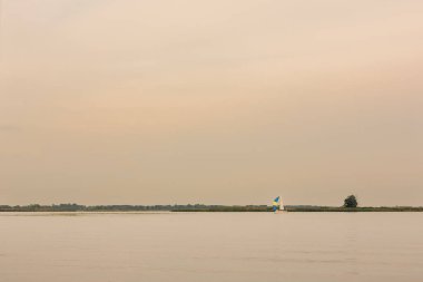 Sakin bir gölde süzülen bir yelkenlinin huzurlu manzarası yumuşak, ılık tonlu gökyüzü ve uzak yeşillik.