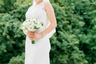 Beyaz dantelli bir gelinlik içinde bir buket beyaz çiçek tutan, yemyeşil bir arka planda duran bir gelin.