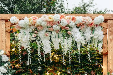 Güzel ahşap düğün kemeri beyaz ve pastel çiçeklerle süslenmiş, mumlar ve ip ışıkları, açık havada romantik bir tören için yemyeşil bir sarmaşığa karşı kurulmuş..