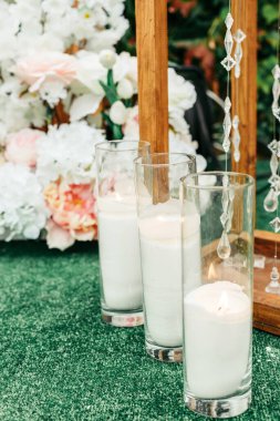 Üç cam mum tutacağı, beyaz mum mumları hafifçe yanıyor, çiçek süslemelerine karşı kurulmuş ve kırsal ahşap çerçeve bir açık hava etkinliğinde.