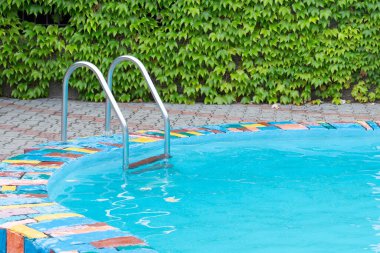 Berrak mavi suyu olan ferahlatıcı bir açık yüzme havuzu, canlı mozaikli bir sınır ve yemyeşil sarmaşık kaplı duvarlarla çevrili..