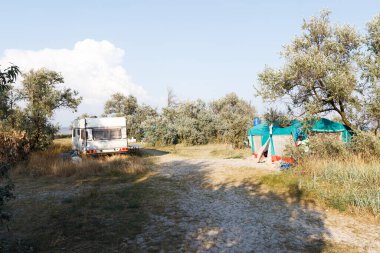 Kırsal bir alanda ağaçlar, çimenler ve açık gökyüzü olan bir karavan ve büyük bir çadır içeren huzurlu bir kamp alanı..