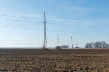 Elektrik direklerinin ve transmisyon kulelerinin bulunduğu bir kırsal alan açık mavi gökyüzünün altında uzanan ve tarımın altyapısını sembolize eden bir tarla..