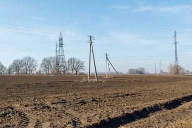 Elektrik direklerinin ve transmisyon kulelerinin bulunduğu bir kırsal alan açık mavi gökyüzünün altında uzanan ve tarımın altyapısını sembolize eden bir tarla..