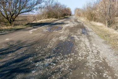 Temiz bir gökyüzünün altında kurumuş çimenler ve çıplak ağaçlarla çevrili büyük çukurları olan hasarlı bir kırsal toprak yol..