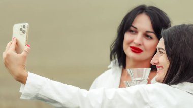 Beyazlar içinde iki şık kadın ellerinde kristal bardaklarla akıllı telefonlarıyla selfie çekiyorlar ve dışarıda sosyal bir toplantının keyfini çıkarıyorlar..