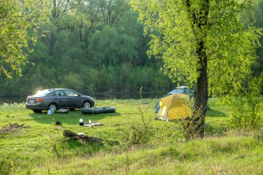 Nehir kıyısında huzurlu bir kamp yeri. İçinde sarı bir çadır, park edilmiş bir araba, bir tekne ve doğada kamp malzemeleri var..