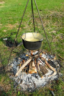 Siyah metal bir tencere tripodun üzerinde açık bir ateşin üzerinde duruyor, yeşil çimlerin üzerinde kırsal bir kamp ortamında yemek pişiriyor..