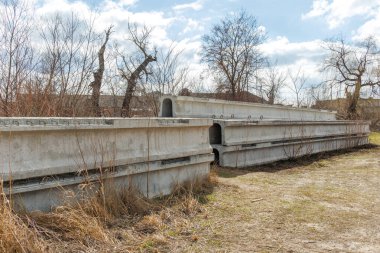 Kuru çimenler ve parçalı bulutlu bir gökyüzünün altında yapraksız ağaçlarla çevrili kırsal bir alanda depolanmış önceden yapılmış beton köprü kirişleri..