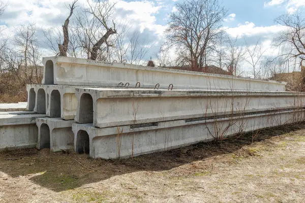 Kuru çimenler ve parçalı bulutlu bir gökyüzünün altında yapraksız ağaçlarla çevrili kırsal bir alanda depolanmış önceden yapılmış beton köprü kirişleri..