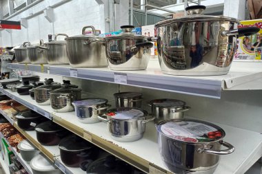 Bir mağaza koridorundaki raflarda düzgünce sıralanmış paslanmaz çelik tencere ve tavalar. Perakende satış ortamında ambalajın canlı renklerini yansıtan parlak tencere ürünleri