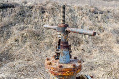 Yıpranmış ve paslı sarı bir gaz boru hattı vanası kuru bir çimenlik alanda duruyor. Yaşlanma, aşınma ve ihmal belirtileri gösteriyor..