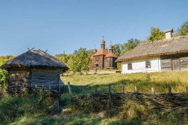Sazdan çatılı evlerin, altın kubbeli ahşap bir kilisenin ve açık mavi gökyüzünün altında örülmüş bir çitin olduğu bir köy manzarası..