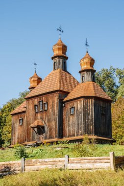 Üç soğan kubbesi olan tarihi ahşap bir kilise. Berrak bir mavi gökyüzünün altında yemyeşil ve kır çitleriyle çevrili..