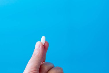 Sağlık ve tıbbı sembolize eden canlı mavi bir arka planı olan tek bir beyaz tablet ya da kapsülü tutan bir insan elinin yakın çekimi.