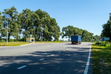 Güneşli kırsal bir yolda giden mavi kamyonet, güneş enerjili sokak lambaları ve yeşil çevresiyle küçük bir otobüs durağından geçiyor..
