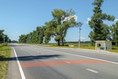 Güneş enerjisiyle çalışan sokak lambaları ve açık mavi bir gökyüzünün altında küçük modern bir otobüs durağı olan boş bir kırsal karayolu..