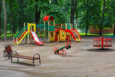 Slaytlar, salıncaklar ve banklarla dolu hareketli oyun alanı yemyeşil bir parkta boş duruyor. Huzurlu ama davetkar bir atmosfer yaratıyor..