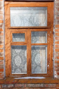 Eski ahşap çerçeveli pencereye yakın çekim, boyası soyulmuş, dantel perdesi ve perdesi karla kaplı, tuğladan bir duvarda..
