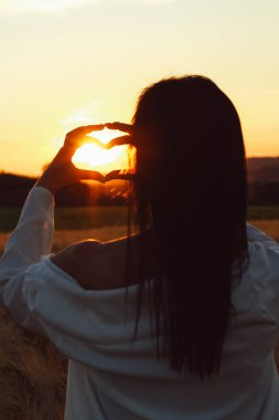 Beyaz gömlekli bir kadının arka görünüşü batan güneşin etrafında elleri ile kalp şekli oluşturan bir kadın..