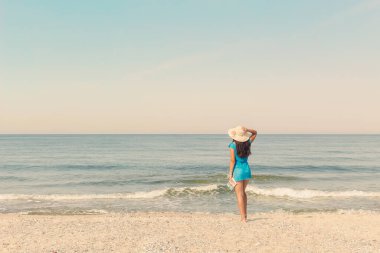 Mavi elbiseli ve güneş şapkalı bir kadının kumlu bir sahilde çıplak ayakla durduğu sakin denizi seyrettiği arka plan..