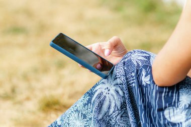 Bir kadının elinde mavi çantalı bir akıllı telefon tutarken, dışarıda otururken elbisesinin üzerinde dinlenirken..