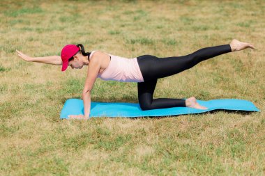 Kuş köpek yogasında dengede duran aktif bir kadın çimenli bir arazide mavi bir mindere poz verip güç ve istikrara odaklanıyor..