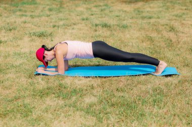 Spor kıyafetli genç bir kadın çimenli bir alanda mavi yoga minderiyle tahta egzersizi yapıyor. Fitness ve sağlıklı yaşam tarzı konsepti.