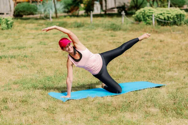 Spor kıyafetli ve kırmızı şapkalı gülümseyen kadın dışarıda yoga yapıyor, bir dizinin üzerinde dengede duruyor, kolları açık ve bacakları havada..