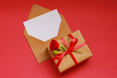 Kırmızı kurdele ve güllerle süslenmiş kraft kağıdına sarılı bir doğum günü hediyesi. Kırmızı arka planda boş bir zarf var. Kutlamayı ve tüm kalbimle dileklerimi simgeliyor..
