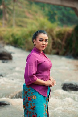 Nehrin önünde duran Asyalı kadın mor bir elbise giyiyordu. Arka planda güzel bir makyaj ve nehir vardı.