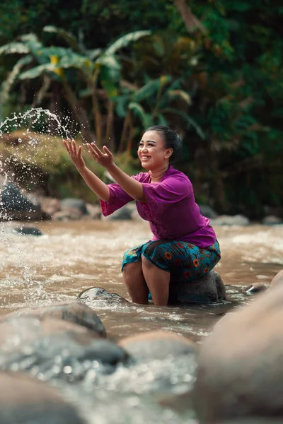 Asian woman playing with dirty water from a dirty river while wearing a purple dress and green skirt near the river