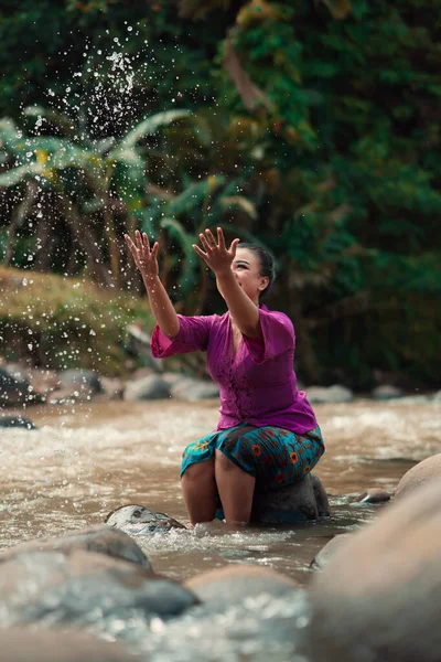 Asian woman playing with dirty water from a dirty river while wearing a purple dress and green skirt near the river