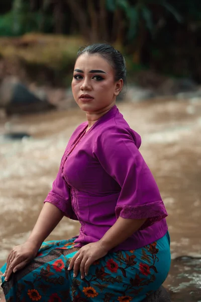 村の中に紫のドレスを着ている間 笑顔で川の近くの岩の上に座って美しいアジアの女性 — ストック写真