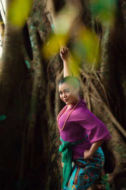 Asyalı bir kadın mor bir elbise ve yüzünde güzel bir gülümsemeyle ormanda dans eder.