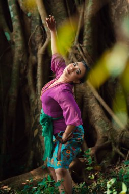 Asyalı bir kadın mor bir elbise ve yüzünde güzel bir gülümsemeyle ormanda dans eder.
