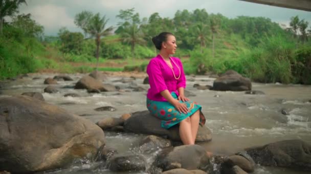 一位身穿传统粉红裙子和绿色裙子的亚洲女人 早上坐在河边的石子上 一边欣赏着村里的美景 — 图库视频影像