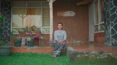 Asyalı bir kadın, sabahları evinin önünde oturmuş gri bir elbise ve siyah bir etek giyerek köyün tadını çıkarıyor.