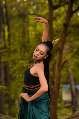 A brave Asian woman in makeup wearing a green costume and gold jewelry with the woods in the background during the morning