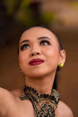 Close-up face of an Asian woman in makeup with red lips and a golden black necklace inside the forest