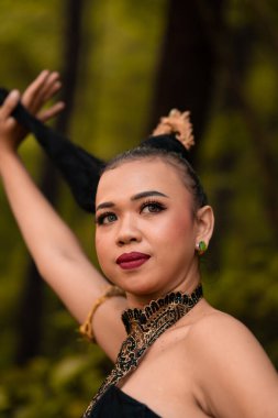Asian woman holding her black hair in a green costume while wearing makeup and posing in front of the forest during the dance festival inside the village