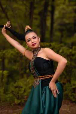 Asian woman holding her black hair in a green costume while wearing makeup and posing in front of the forest during the dance festival inside the village
