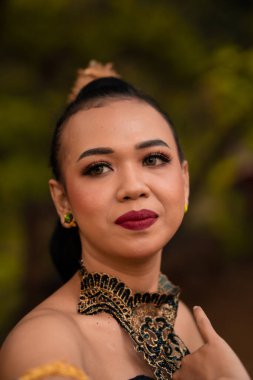Close-up face of an Asian woman in makeup with red lips and a golden black necklace inside the forest