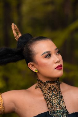 Gorgeous Asian woman in makeup with black hair and red lipstick wearing golden jewelry during the dance performance in front of the jungle