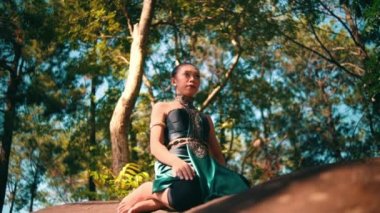 A beautiful Asian woman in a green costume sitting on a big rock while visiting the forest lot of trees inside the village