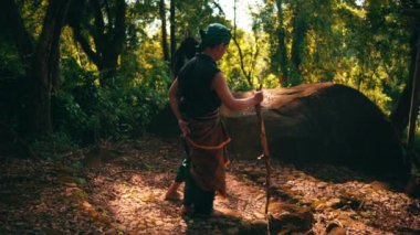 An Asian couple walking together in traditional green costumes inside the forest while the man holding a stick beside the woman during the morning
