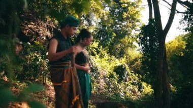 An Asian couple walking together in traditional green costumes inside the forest while the man holding a stick beside the woman during the morning